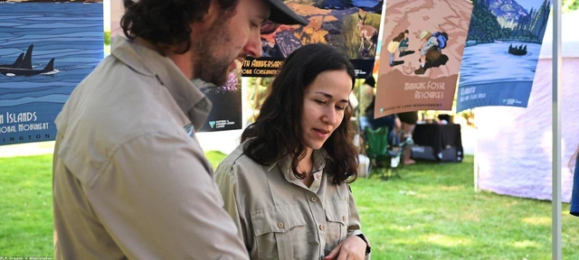 Dos compañeros con camisa caqui revisan algo juntos en un estand al aire libre con carteles